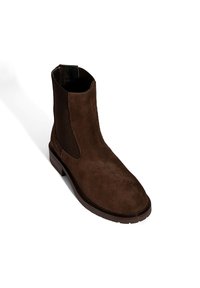 Bottine en daim marron avec des panneaux latéraux élastiques et une semelle plate robuste. Présente une texture lisse et un design de bout légèrement pointu.