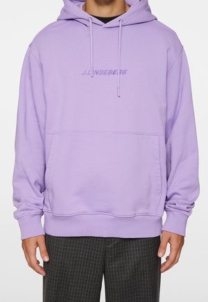 Osoba v světle fialové mikině s kapucí a přední kapsou na břiše s logem "J. Lindeberg" na hrudi, v kombinaci s tmavými kostkovanými kalhotami.
