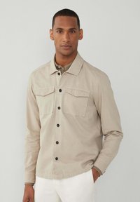 Hellbeige Button-Up-Hemd mit zwei Brusttaschen, schwarzen Knöpfen und einem Kragen. Lange Ärmel mit glatter Textur.