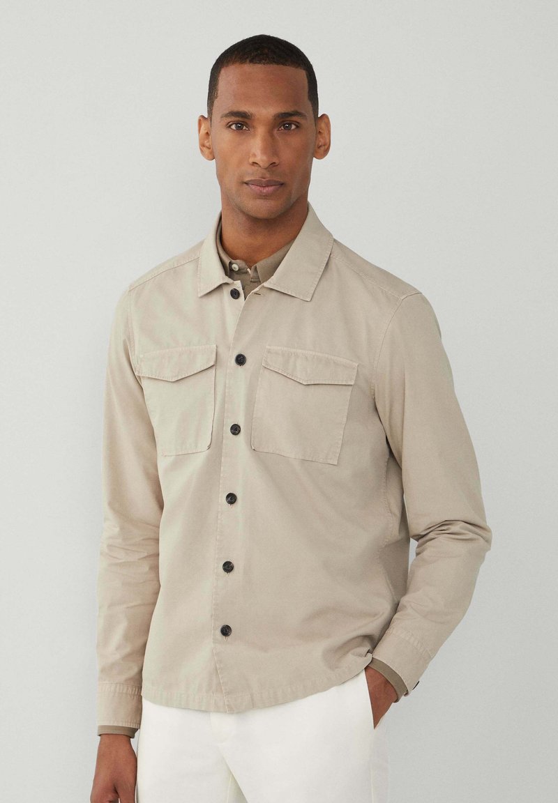 Hellbeige Button-Up-Hemd mit zwei Brusttaschen, schwarzen Knöpfen und einem Kragen. Lange Ärmel mit glatter Textur.