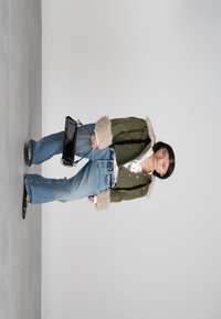 Mulher vestindo um casaco de pelúcia verde, camisa branca, calças jeans largas azuis, sapatos pretos, e segurando uma pequena bolsa preta, em pé contra um fundo simples.