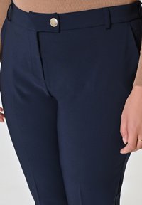 Pantaloni tailored blu navy, realizzati in tessuto liscio, con chiusura frontale a bottone e tasche laterali, che presentano una silhouette slim.