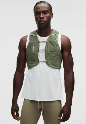 Hombre atlético con cabello trenzado, vistiendo una camiseta blanca sin mangas, chaleco verde oliva con cremallera y pantalones beige con cordones.