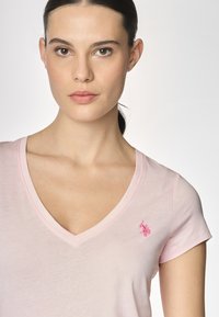 T-shirt a V collo rosa chiaro realizzato in tessuto morbido, con un piccolo logo ricamato sulla parte sinistra del petto. Maniche corte.