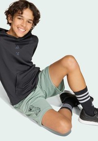 Usmievajúci sa chlapec sedí na podlahe, má na sebe čiernu mikinu, svetlozelené šortky s pixelovým vzorom, čierne tenisky a čierne ponožky s bielymi pruhmi.