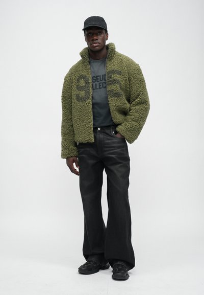 Fuzzy olive green jacket with high collar, black graphic t-shirt, wide-leg black jeans, and black shoes; overall relaxed fit.