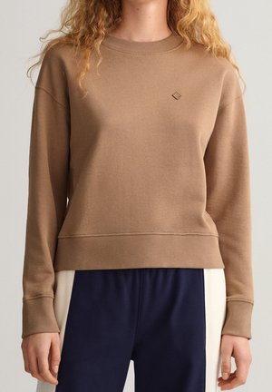 Vrouw draagt een bruin sweatshirt met lange mouwen, geribde manchetten en zoom, gecombineerd met marineblauwe broek met witte zijpanelen.