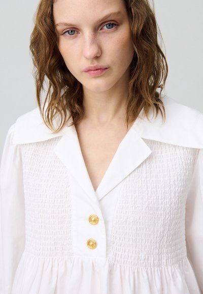 Vestido blanco con cuerpo fruncido, gran cuello y botones dorados. Presenta una falda acampanada y una textura de tela suave.