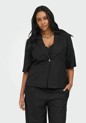 Femme aux longs cheveux noirs porte un blazer noir à manches courtes, un pantalon assorti, un collier chaîne en or et des boucles d'oreilles créoles, debout avec une main dans la poche.