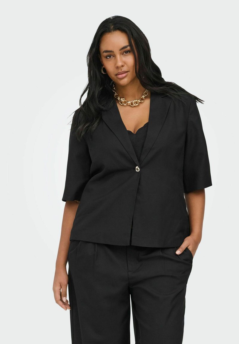 Femme aux longs cheveux noirs porte un blazer noir à manches courtes, un pantalon assorti, un collier chaîne en or et des boucles d'oreilles créoles, debout avec une main dans la poche.