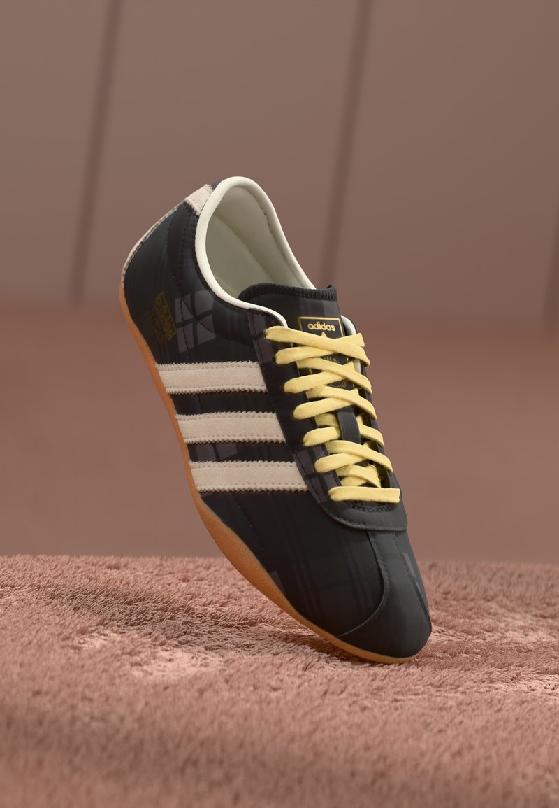 Sapatilhas pretas de desporto com três riscas brancas, atacadores amarelos e um acabamento texturizado. Apresentam um design curvo e sola de borracha.