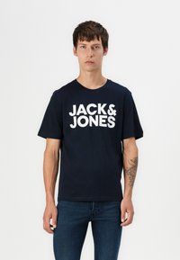 Tamsiai mėlyna medvilninė marškinėlių su trumpomis rankovėmis, su dideliu baltu "JACK & JONES" logotipu priekyje. Klasikinio apvalaus kaklo dizainas.