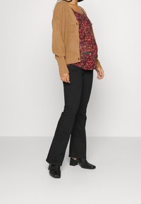 Cardigan beige lavorato a maglia, top con motivo floreale nei toni del rosso, nero e viola, abbinato a pantaloni neri a campana e scarpe nere con tacco.