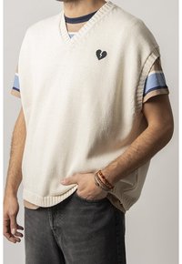 Cremfarbener gestrickter Pullover mit V-Ausschnitt, ausgestattet mit einem schwarzen Herz-Logo. Über einem gestreiften Hemd geschichtet, aus strukturiertem Stoff und im entspannten Schnitt.