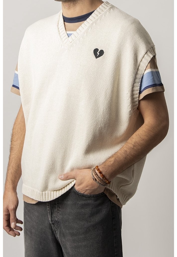 Cremfarbener gestrickter Pullover mit V-Ausschnitt, ausgestattet mit einem schwarzen Herz-Logo. Über einem gestreiften Hemd geschichtet, aus strukturiertem Stoff und im entspannten Schnitt.