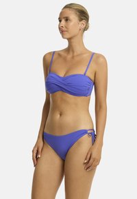 Model in einem einfarbigen lila Bikini mit einem verdrehten Oberteil und tief geschnittenen Höschen, die mit leichtem Profil und natürlichem Make-up vor einem weißen Hintergrund steht.