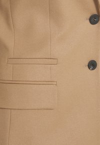 Blazer doppiopetto color tan realizzato in tessuto testurizzato, con due bottoni neri e una tasca a filetto sul lato sinistro.