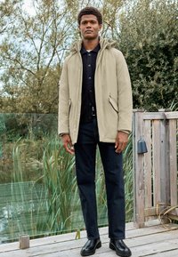 Beige Parka-Jacke mit Kapuze, Frontreißverschluss und seitlichen Taschen, über einem schwarzen Strickshirt getragen. Dazu dunkle Jeans und schwarze Stiefel.