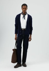 Marineblauer Strickcardigan mit Kabelmuster, weißes T-Shirt, dunkelblaue Jeans, braune Loafers. Braune Tragetasche, blau gemusterter Schal am Hals gebunden.