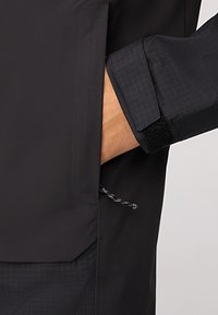 Chaqueta negra con un patrón texturizado, puños ajustables con velcro y un bolsillo lateral con un detalle de cordón.