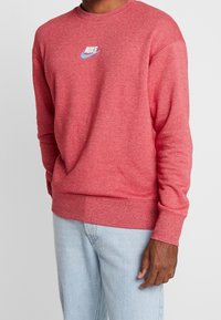 Rotes Sweatshirt mit Rundhalsausschnitt aus weichem Stoff und geripptem Saum, mit kleinem weißen und hellblauen Nike-Logo auf der Brust.