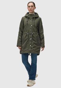Parka impermeable verde con capucha ajustable, botones a presión y dos bolsillos laterales. Lleva puesta sobre unos jeans azules y zapatillas color crema.