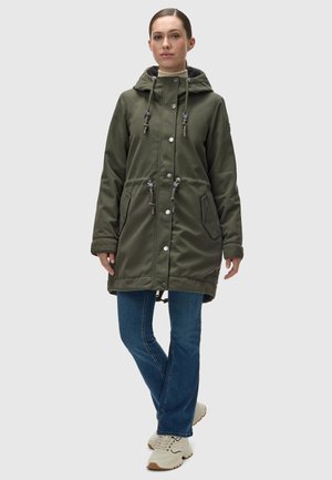 Green waterproof parka with a drawstring hood, snap buttons, and two side pockets. Worn over blue jeans and cream sneakers.