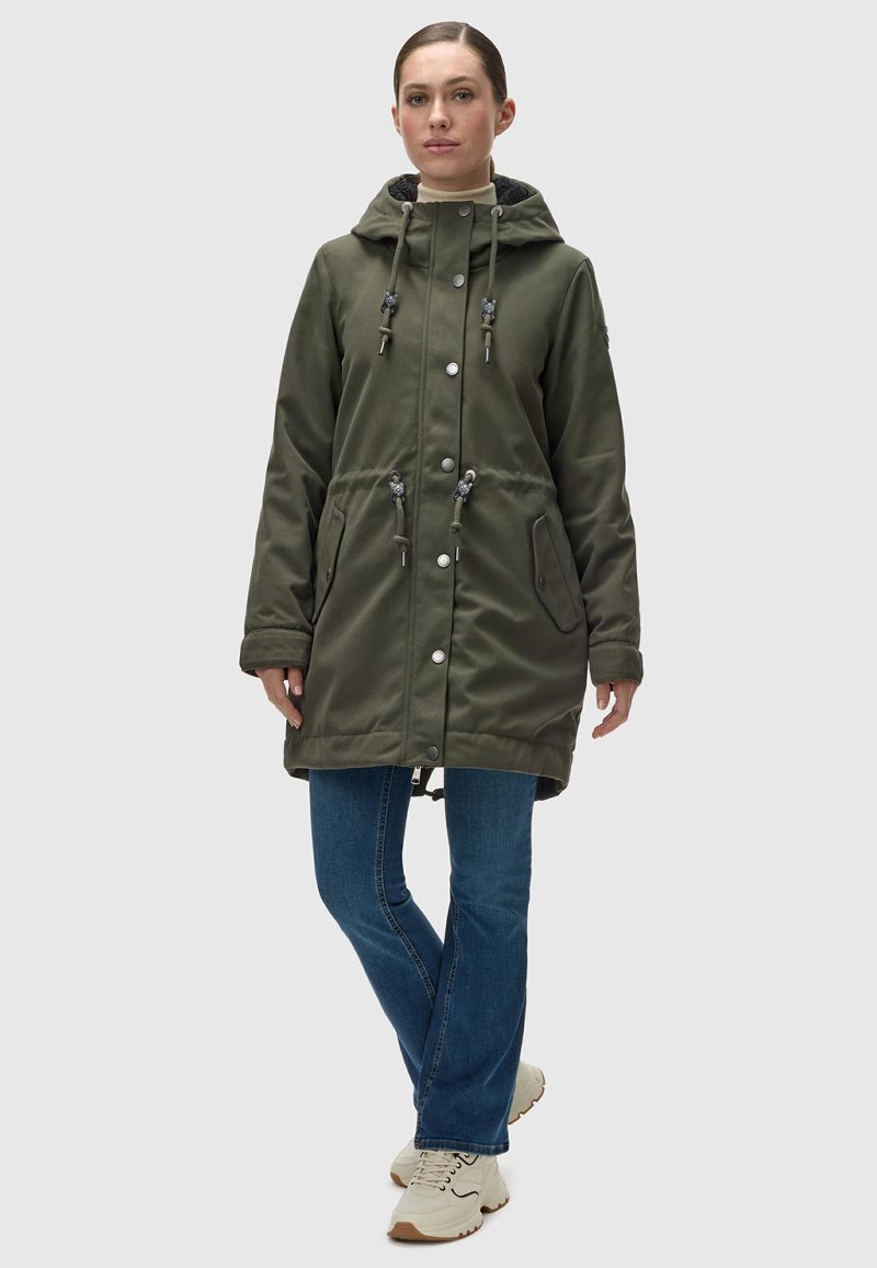 Parka impermeable verde con capucha ajustable, botones a presión y dos bolsillos laterales. Lleva puesta sobre unos jeans azules y zapatillas color crema.