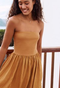 Váll nélküli ruha meleg, mustár színben, texturált, testhez simuló bodéval és lágyan gyűrt szoknyával. Arany nyaklánccal díszítve.