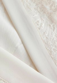 Tessuto bianco con trame diverse, includendo un'area liscia e una sezione con un motivo soffice e frangiato. Aspetto morbido e leggero.