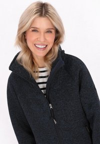 Mustat ja siniset fleece-takit, joissa on korkea kaulus ja vetoketjukiinnitys, sekä rakenteellinen malli ja vetoketjullinen rintatasku.