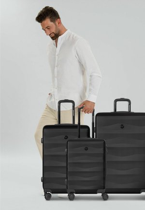 Uomo con camicia bianca e pantaloni beige in piedi vicino a tre valigie nere di diverse dimensioni con manici estesi.