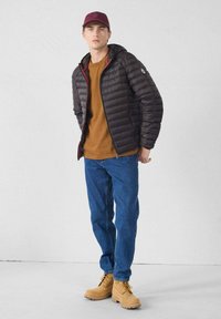 Giovane uomo con cappellino color bordeaux, giacca imbottita nera, camicia color senape, jeans blu e stivali da lavoro color cuoio, in piedi contro un muro bianco.