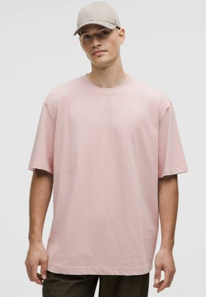 T-shirt en coton rose surdimensionné avec des manches courtes et un col rond, accompagné d'une casquette beige clair. Design minimaliste, texture lisse.