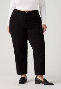 Jeans de mezclilla negra de cintura alta con corte recto, costuras visibles, bolsillos delanteros y cierre de botones. Combinados con mocasines negros.