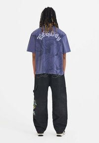 Lilla grafisk t-shirt med et stort design på ryggen, parret med løstsiddende sorte cargo-bukser udsmykket med farverig broderi.