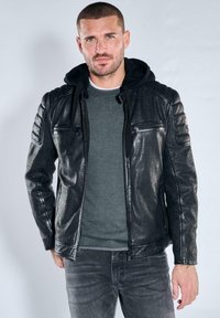 Mann mit kurzen Haaren und Bart, der eine schwarze Lederjacke mit gesteppter Schulterpartie und einen grauen Pullover trägt, vor einem einfarbigen Hintergrund steht.
