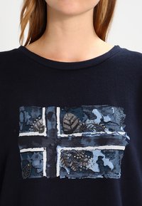 Mujer que lleva una camisa azul oscuro con un diseño texturizado y con cuentas de la bandera de Noruega en el pecho, que presenta colores azul, blanco y negro.