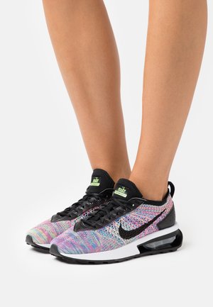 Nohy v barevných pletených teniskách Nike s černými tkaničkami, černým logem Nike, bílou podešví a viditelným vzduchovým polštářem na bílém pozadí.