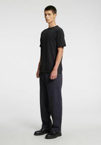 Juoda medvilninė marškinėliai su apvaliu kaklu, laisvo kirpimo, derinami su tamsiai mėlynomis kelnėmis ir juodais odiniais batais, stovintys priešais paprastą foną.