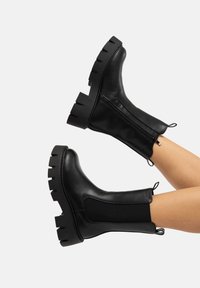 Schwarze Knöchelstiefel aus glattem Leder mit einer klobigen, strukturierten Sohle, seitlichem Reißverschluss und elastischem Einsatz für eine bessere Passform.