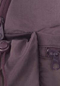 Szczegółowy widok fioletowego plecaka z materiału, ukazujący kieszeń na zamek błyskawiczny z uchwytem, elastyczny sznurek ściągający oraz teksturowany materiał dla zwiększonej trwałości.