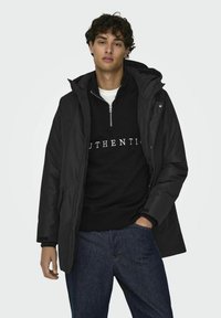 Chaqueta negra con capucha de textura suave, llevada sobre un sudadera negra con texto blanco. El modelo pose con las manos en los bolsillos, vistiendo unos jeans.