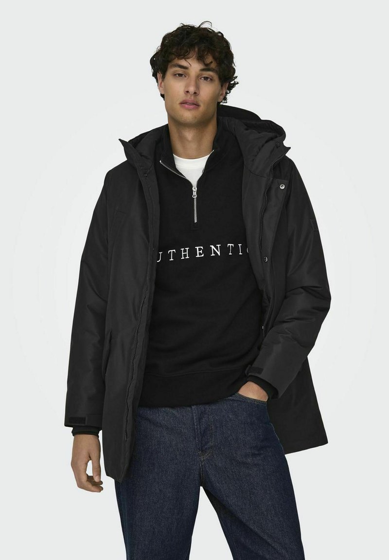 Chaqueta negra con capucha de textura suave, llevada sobre un sudadera negra con texto blanco. El modelo pose con las manos en los bolsillos, vistiendo unos jeans.