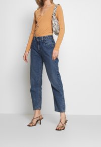 Beige Langarm-Wickeloberteil, kombiniert mit hoch taillierten blauen Jeans. Akzentuiert mit einer Tasche im Schlangenledermuster und Riemchen-Heels.