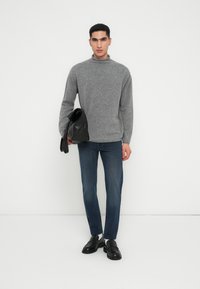 Pulôver cinza com gola alta, jeans azul escuro e bolsa de couro preta. Modelo veste sapatos loafers pretos, apresentando linhas limpas e um design moderno.