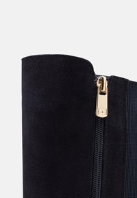 Mörkblå mockaboot med en slät yta, med en guld dragkedja på sidan och en elastisk panel för stretching.