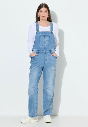 Giovane donna in piedi contro una parete chiara, indossa una salopette di jeans azzurro chiaro, una maglia a maniche lunghe bianca, sneakers bianche, con le mani nelle tasche, espressione neutra.