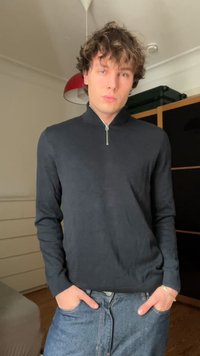 Pull noir zippé avec des manches longues, fait d'un tissu lisse. Associé à un jean en denim bleu, les mains dans les poches, debout dans une pièce.