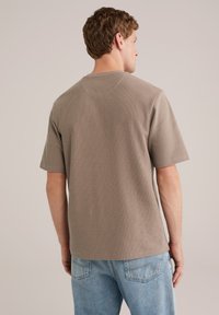 Rjava kratka majica s kratkimi rokavi iz teksturirane tkanine; ima krojeni izrez in sproščen kroj, v kombinaciji s svetlo modrimi džinsami.